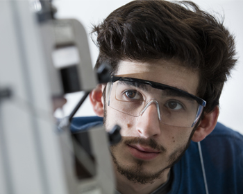 A student in safety glasses closely inspecting a piece of equipment