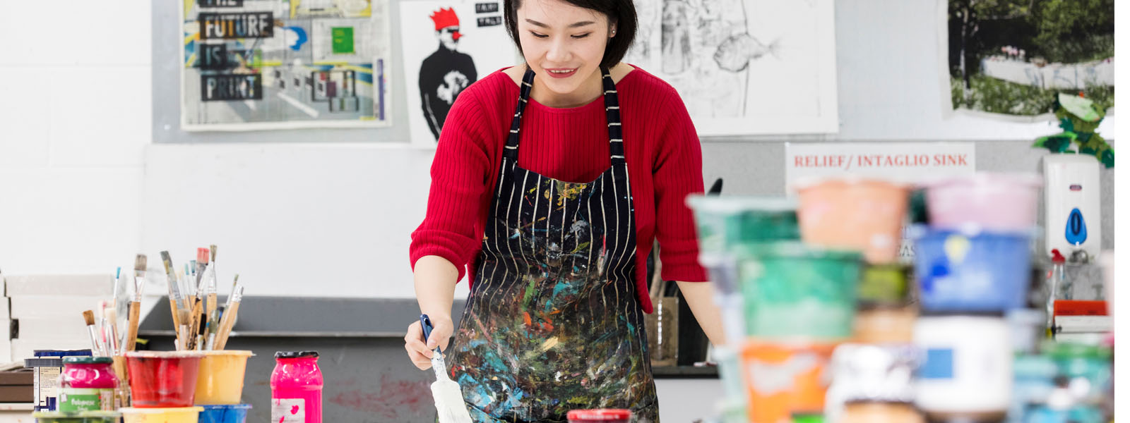 Female student painting in the studio with colourful paint bottles surrounding her