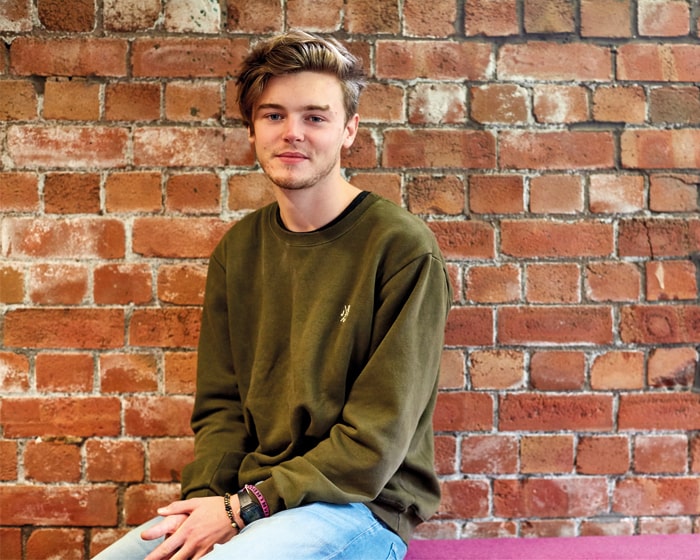Student sat on the arm of a pink sofa with an exposed brick wall in the background