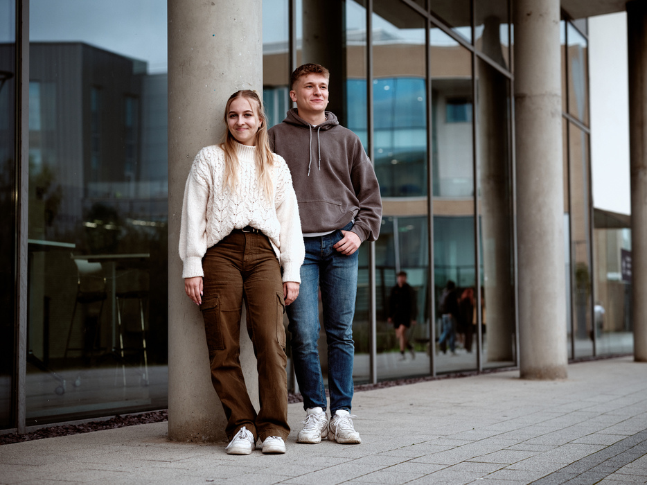 Two students stood against a concrete pillar outside the Nicola de la Haye Building