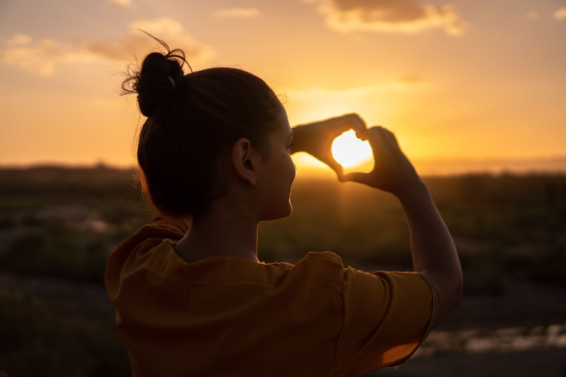 Woman making a heart shape with both hands facing a sunset