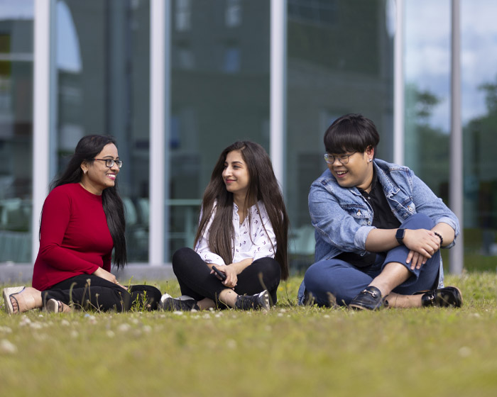 A group of students sitting and talking on the grass in front of a glass building