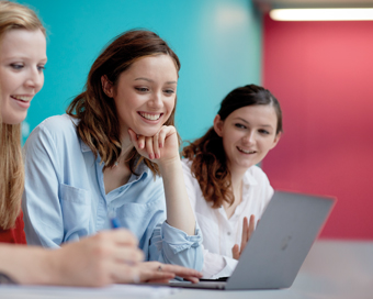 Three ladies sat at a desk, looking at a laptop and smiling