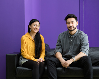 Female student in yellow top and male student in grey shirt sat on a black sofa, with a purple wall in the background