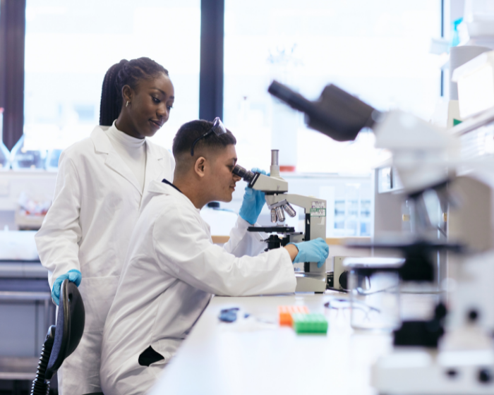 Two students in lab coats in a lab, with one sat at a desk using a microscope, and the other stood behind