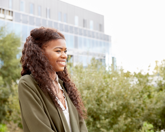 A female student stood outside the Isaac Newton Building with greenery in the background