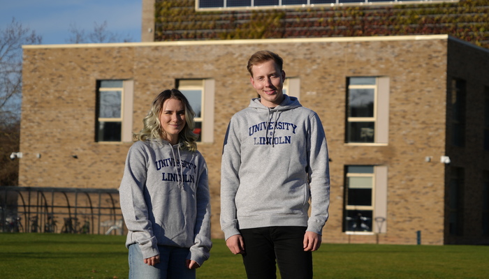 A male and a female student both wearing grey branded University of Lincoln hoodies stood outside the Ross Lucas Medical Sciences Building
