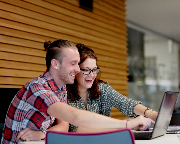 Two student sat at a table in the library, pointing at a laptop screen and smiling