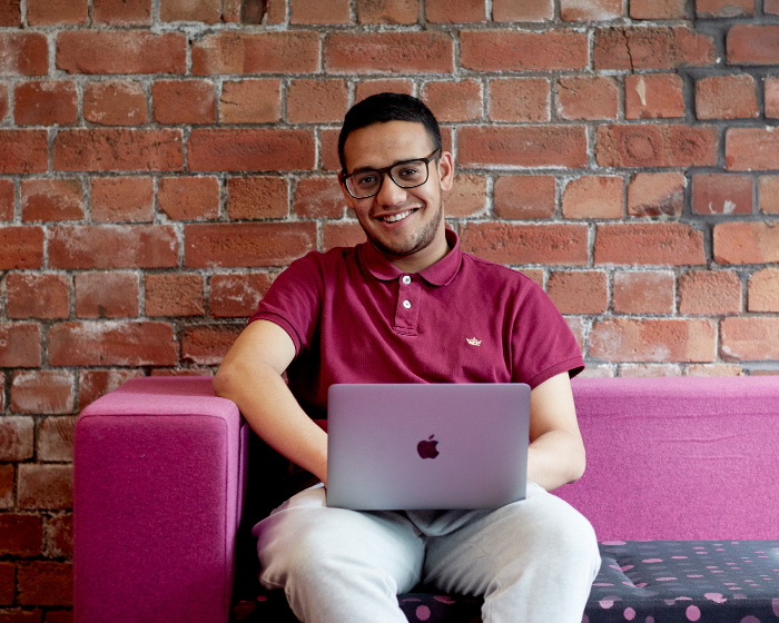 Student with a laptop on his lap, sat on a pink sofa with an exposed brick wall behind him