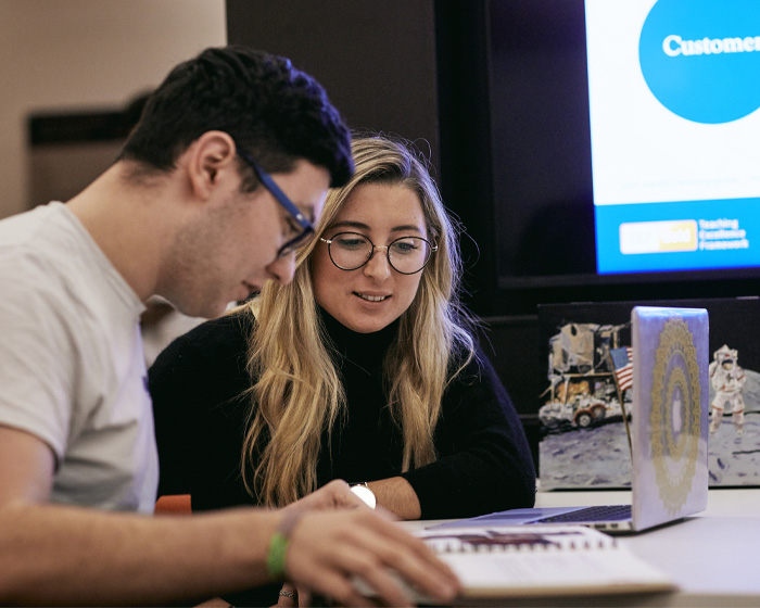 Two students sat at a desk reading and working on a laptop